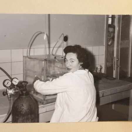 Black and white image of women sitting at work in a laboratory and wearing a white lab coat turning to look at the camera.